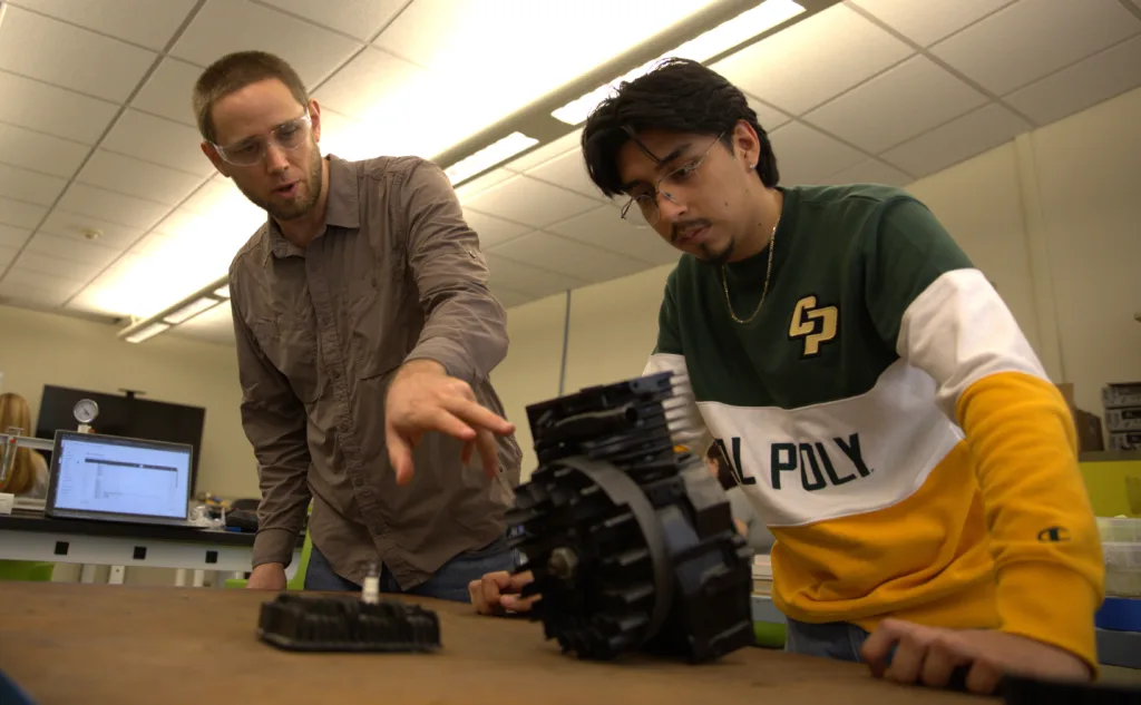 A college lecturer points something out to a student working on a small engine