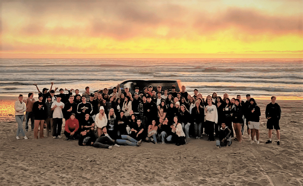 he Mustang Surf Club at a bonfire on the beach at sunset.