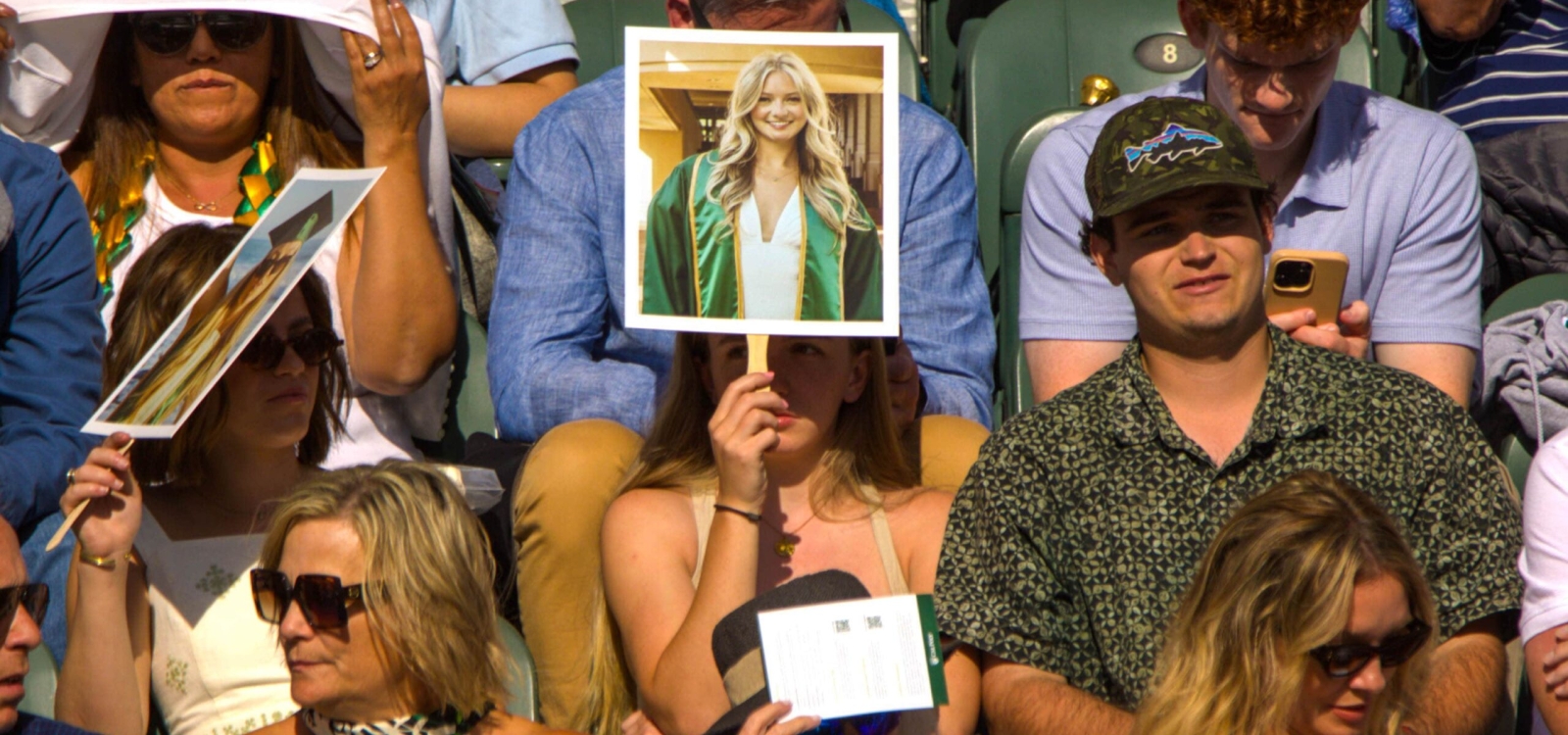An attendee in the stands holding up a photo of a graduate