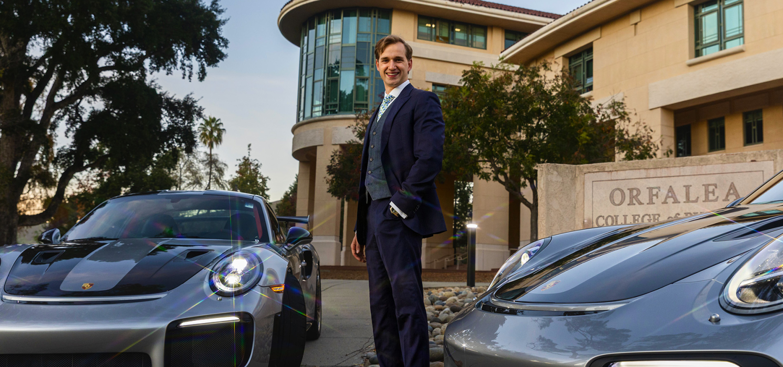 Alumni poses with two Porsches outside business building