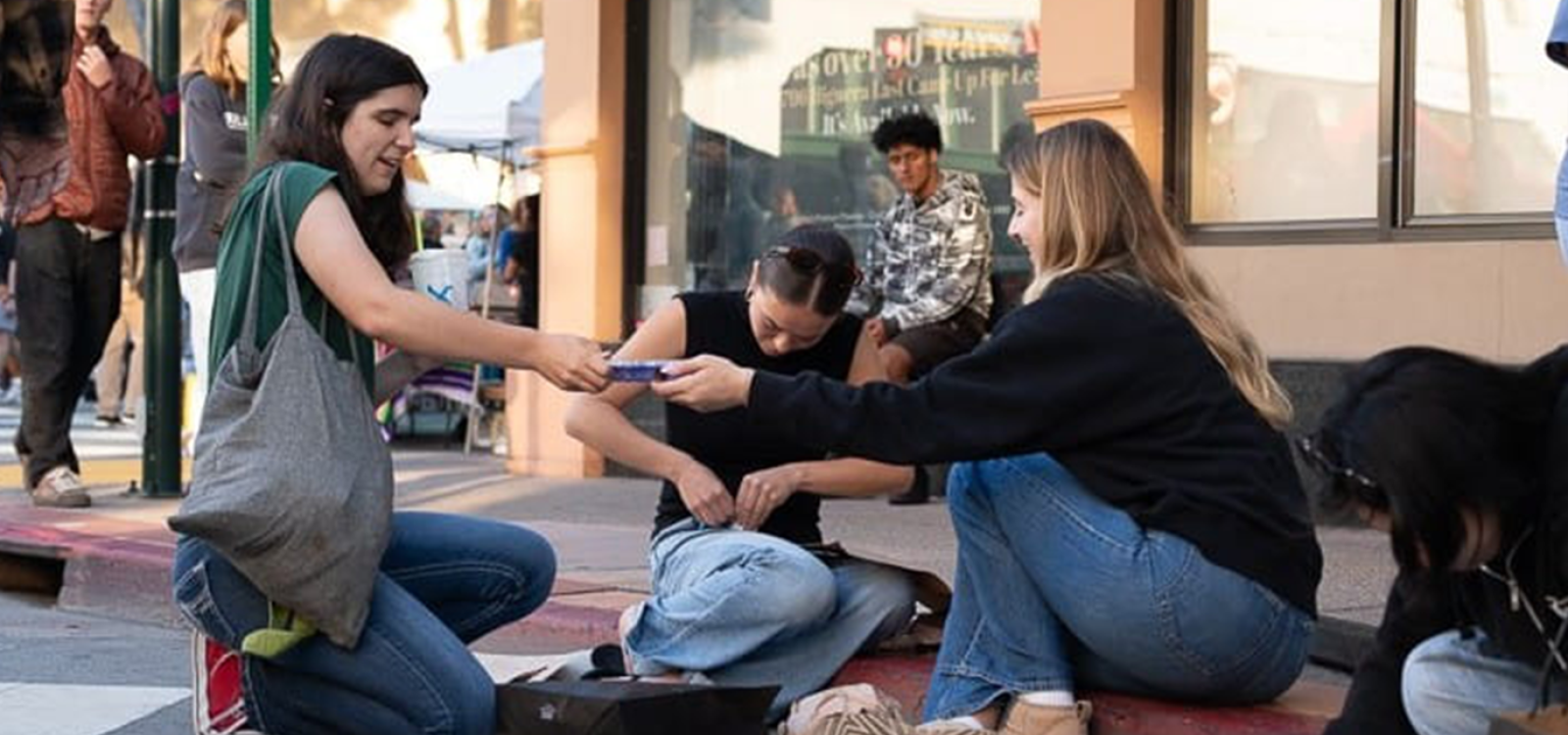 Three students create an egg drop package while sitting on a curb