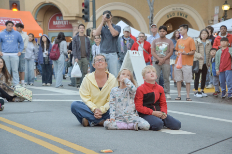 Members of the audience, sitting on the ground, look up to the egg drop