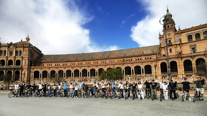 A large group of people with bicycles pose together in a plaza in front of a historic building with arched walkways and towers under a partly cloudy sky.